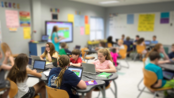 Diverse students in a class work on a project while teacher provides instructional using a Lightspeed Classroom Audio System